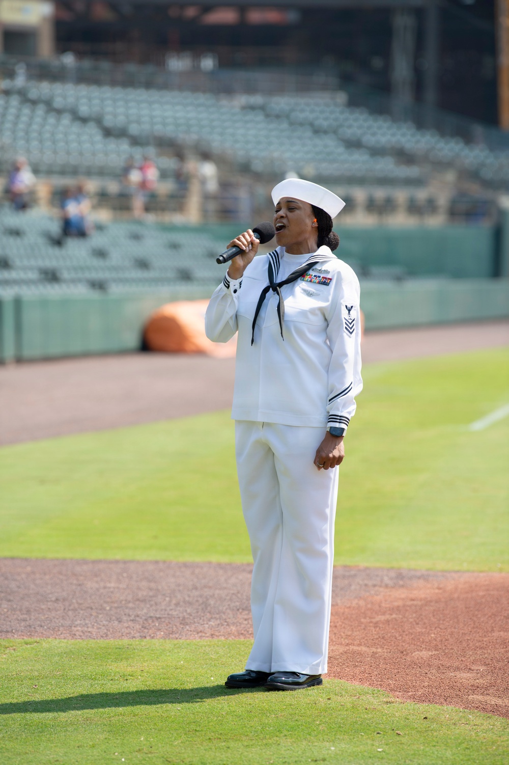 A U.S. Navy sailor in dress white uniform is singing into a handheld microphone on the field of a baseball stadium. The sailor is wearing a Crackerjack-style uniform with visible rank chevrons, ribbons, and a white dixie cup hat. The stadium seats are largely empty, suggesting this is a pre-game ceremony, likely a national anthem performance.
