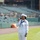 A U.S. Navy sailor in dress white uniform is singing into a handheld microphone on the field of a baseball stadium. The sailor is wearing a Crackerjack-style uniform with visible rank chevrons, ribbons, and a white dixie cup hat. The stadium seats are largely empty, suggesting this is a pre-game ceremony, likely a national anthem performance.