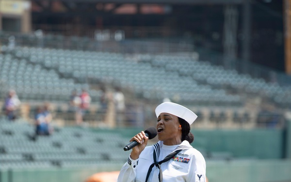 A U.S. Navy sailor in dress white uniform is singing into a handheld microphone on the field of a baseball stadium. The sailor is wearing a Crackerjack-style uniform with visible rank chevrons, ribbons, and a white dixie cup hat. The stadium seats are largely empty, suggesting this is a pre-game ceremony, likely a national anthem performance.