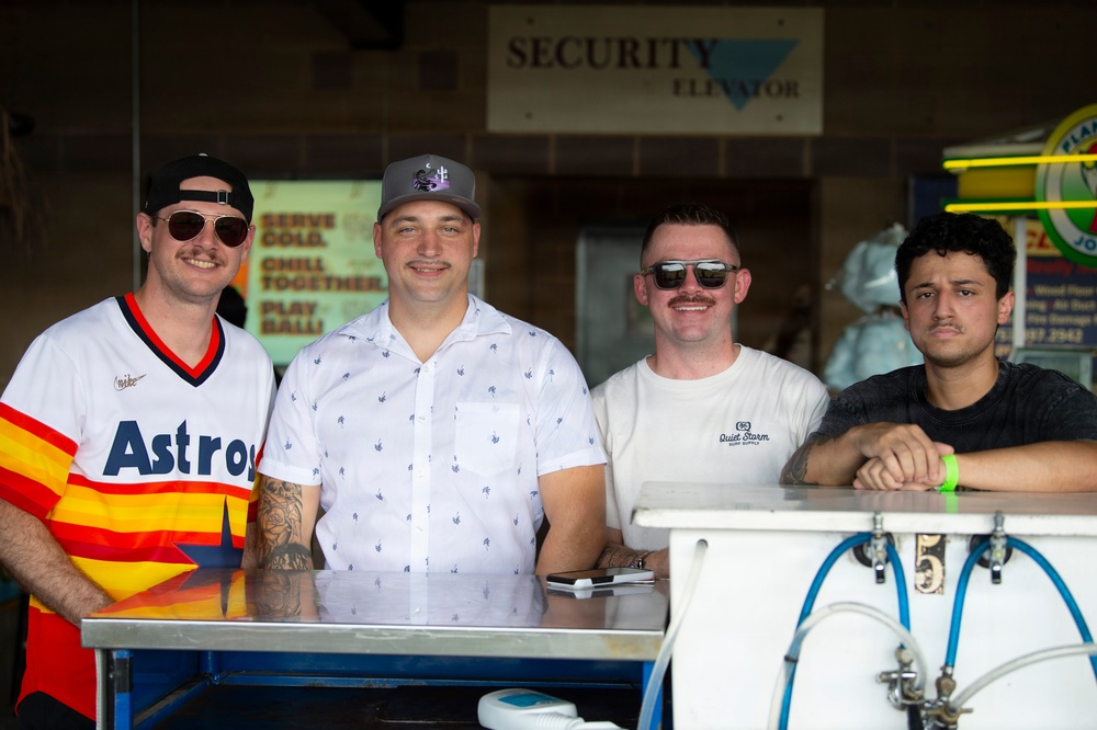 Four men are posing together behind a counter or bar setup, likely at a stadium or outdoor venue. The setting appears to be a concession or beverage stand, with beer tap lines visible in the foreground. The group appears relaxed and in good spirits, suggesting a casual or celebratory occasion.