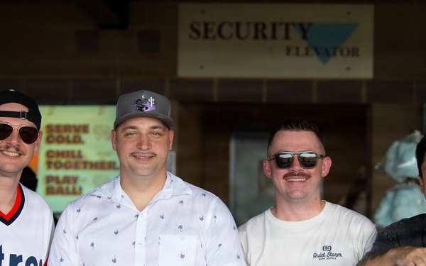 Four men are posing together behind a counter or bar setup, likely at a stadium or outdoor venue. The setting appears to be a concession or beverage stand, with beer tap lines visible in the foreground. The group appears relaxed and in good spirits, suggesting a casual or celebratory occasion.