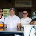 Four men are posing together behind a counter or bar setup, likely at a stadium or outdoor venue. The setting appears to be a concession or beverage stand, with beer tap lines visible in the foreground. The group appears relaxed and in good spirits, suggesting a casual or celebratory occasion.