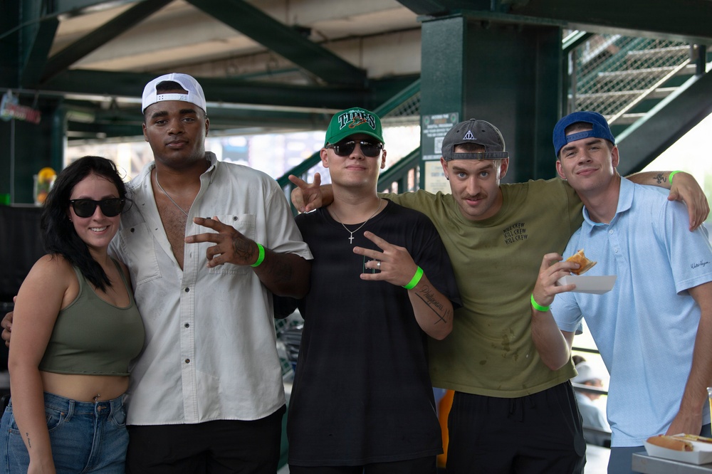 Five young adults pose together for a group photo at what appears to be a sports stadium or arena concourse area. The group consists of one woman and four men, several wearing baseball caps and green wristbands, suggesting they are attending the same event. The atmosphere is casual and social, with the individuals smiling and making hand gestures for the camera.