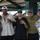 Five young adults pose together for a group photo at what appears to be a sports stadium or arena concourse area. The group consists of one woman and four men, several wearing baseball caps and green wristbands, suggesting they are attending the same event. The atmosphere is casual and social, with the individuals smiling and making hand gestures for the camera.
