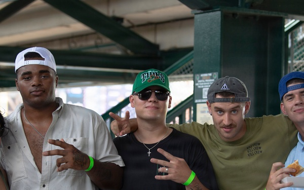 Five young adults pose together for a group photo at what appears to be a sports stadium or arena concourse area. The group consists of one woman and four men, several wearing baseball caps and green wristbands, suggesting they are attending the same event. The atmosphere is casual and social, with the individuals smiling and making hand gestures for the camera.