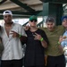 Five young adults pose together for a group photo at what appears to be a sports stadium or arena concourse area. The group consists of one woman and four men, several wearing baseball caps and green wristbands, suggesting they are attending the same event. The atmosphere is casual and social, with the individuals smiling and making hand gestures for the camera.