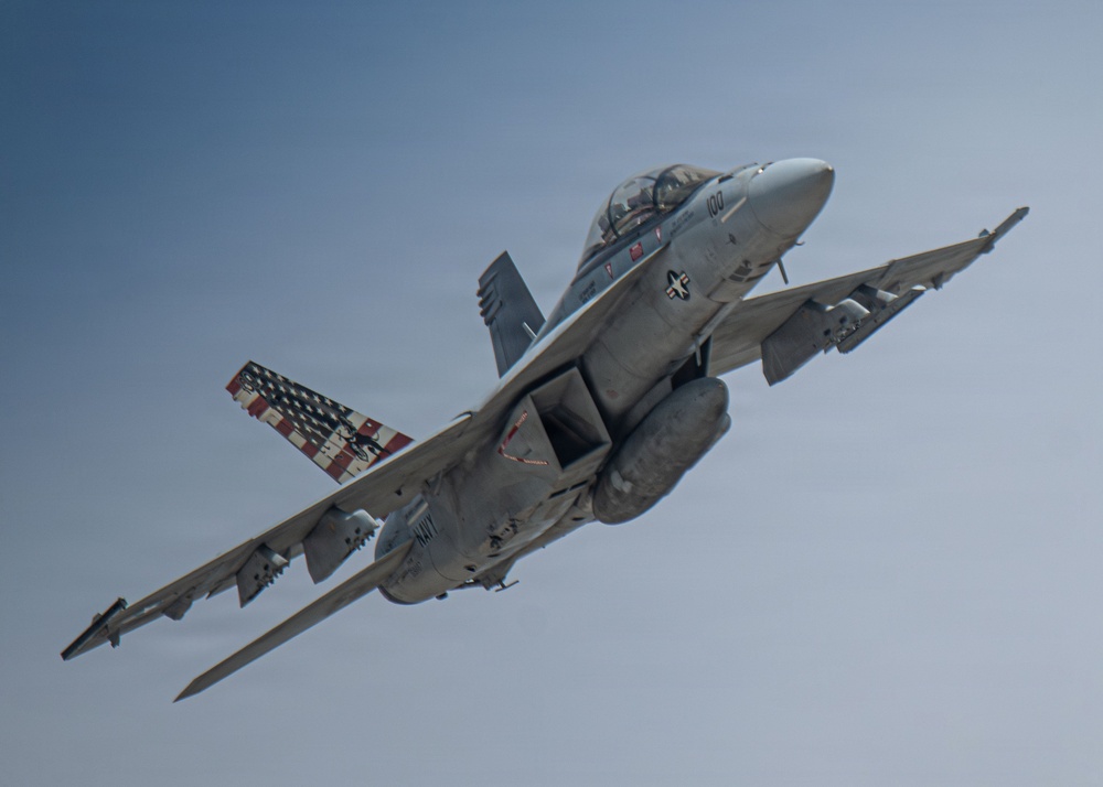 A U.S. Navy tactical jet aircraft, marked with the number '100' on the nose, is captured in flight against a clear blue-gray sky. The aircraft features a distinctive American flag livery on one of its tail fins, along with visible weapons pylons and external fuel tanks. The aircraft appears to be in a climbing or banking maneuver with landing gear retracted.