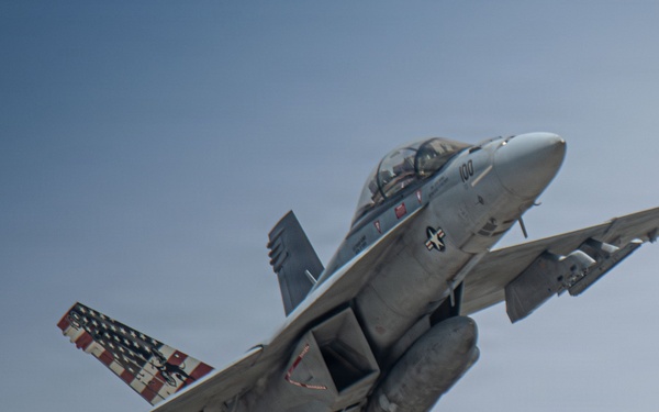 A U.S. Navy tactical jet aircraft, marked with the number '100' on the nose, is captured in flight against a clear blue-gray sky. The aircraft features a distinctive American flag livery on one of its tail fins, along with visible weapons pylons and external fuel tanks. The aircraft appears to be in a climbing or banking maneuver with landing gear retracted.