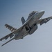 A U.S. Navy tactical jet aircraft, marked with the number '100' on the nose, is captured in flight against a clear blue-gray sky. The aircraft features a distinctive American flag livery on one of its tail fins, along with visible weapons pylons and external fuel tanks. The aircraft appears to be in a climbing or banking maneuver with landing gear retracted.