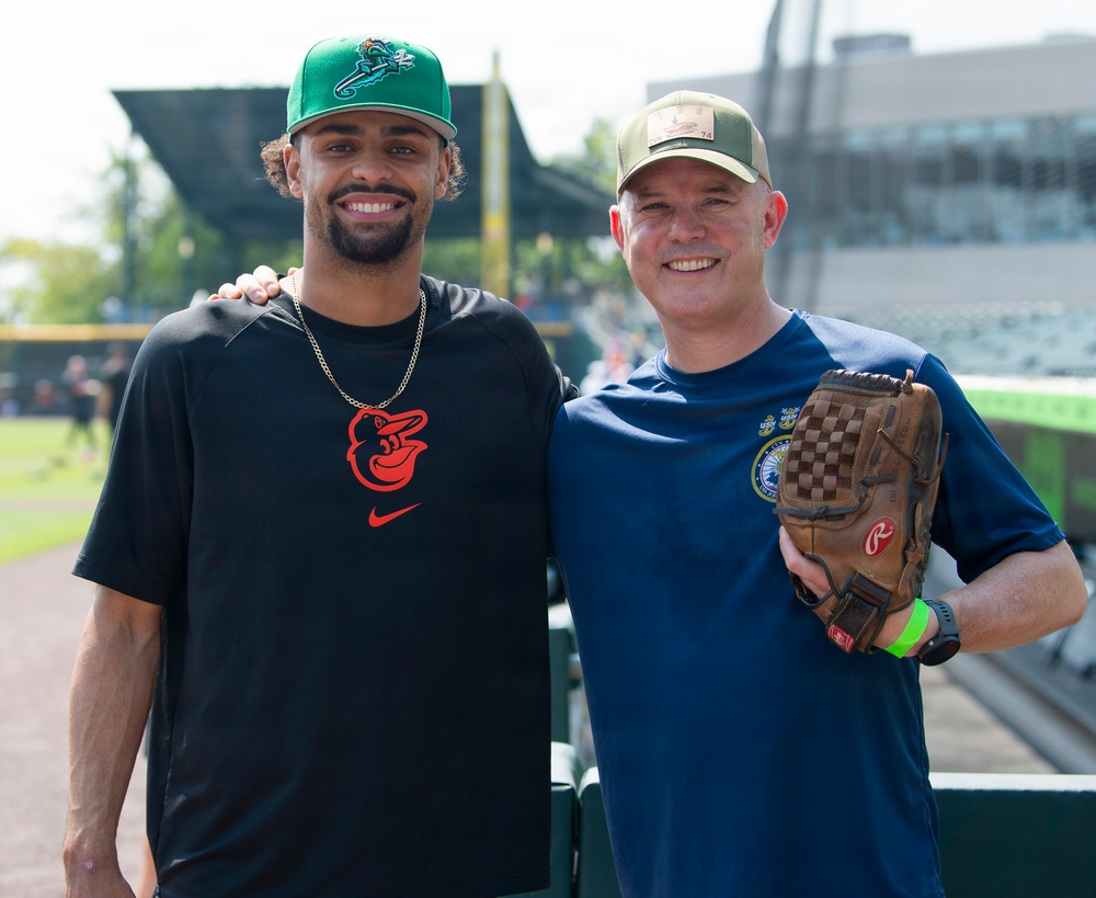 Two men pose together smiling at what appears to be a baseball stadium or training facility. The man on the left wears a Baltimore Orioles Nike shirt and a green baseball cap, while the man on the right holds a Rawlings baseball glove and wears a navy blue shirt with a patch and an olive/tan cap. The setting appears to be near the dugout or field-level area of a ballpark.