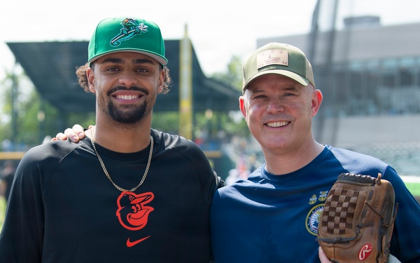 Two men pose together smiling at what appears to be a baseball stadium or training facility. The man on the left wears a Baltimore Orioles Nike shirt and a green baseball cap, while the man on the right holds a Rawlings baseball glove and wears a navy blue shirt with a patch and an olive/tan cap. The setting appears to be near the dugout or field-level area of a ballpark.