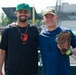 Two men pose together smiling at what appears to be a baseball stadium or training facility. The man on the left wears a Baltimore Orioles Nike shirt and a green baseball cap, while the man on the right holds a Rawlings baseball glove and wears a navy blue shirt with a patch and an olive/tan cap. The setting appears to be near the dugout or field-level area of a ballpark.