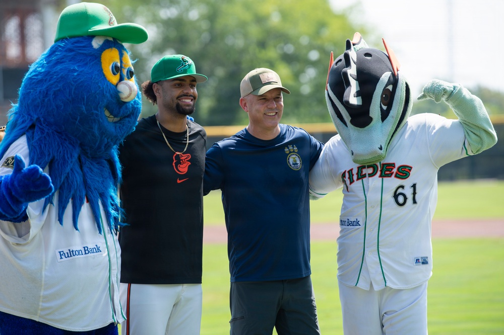 Two men pose for a photo on a baseball field flanked by two team mascots. The left mascot is a large blue furry bird-like creature wearing a green cap with 'Fulton Bank' sponsorship, while the right mascot is a fish/tides-themed character wearing a white uniform with 'Tides' and number 61. The setting appears to be a minor league baseball stadium on a sunny day.