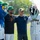 Two men pose for a photo on a baseball field flanked by two team mascots. The left mascot is a large blue furry bird-like creature wearing a green cap with 'Fulton Bank' sponsorship, while the right mascot is a fish/tides-themed character wearing a white uniform with 'Tides' and number 61. The setting appears to be a minor league baseball stadium on a sunny day.