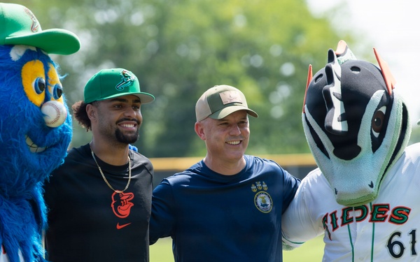 Two men pose for a photo on a baseball field flanked by two team mascots. The left mascot is a large blue furry bird-like creature wearing a green cap with 'Fulton Bank' sponsorship, while the right mascot is a fish/tides-themed character wearing a white uniform with 'Tides' and number 61. The setting appears to be a minor league baseball stadium on a sunny day.