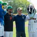 Two men pose for a photo on a baseball field flanked by two team mascots. The left mascot is a large blue furry bird-like creature wearing a green cap with 'Fulton Bank' sponsorship, while the right mascot is a fish/tides-themed character wearing a white uniform with 'Tides' and number 61. The setting appears to be a minor league baseball stadium on a sunny day.