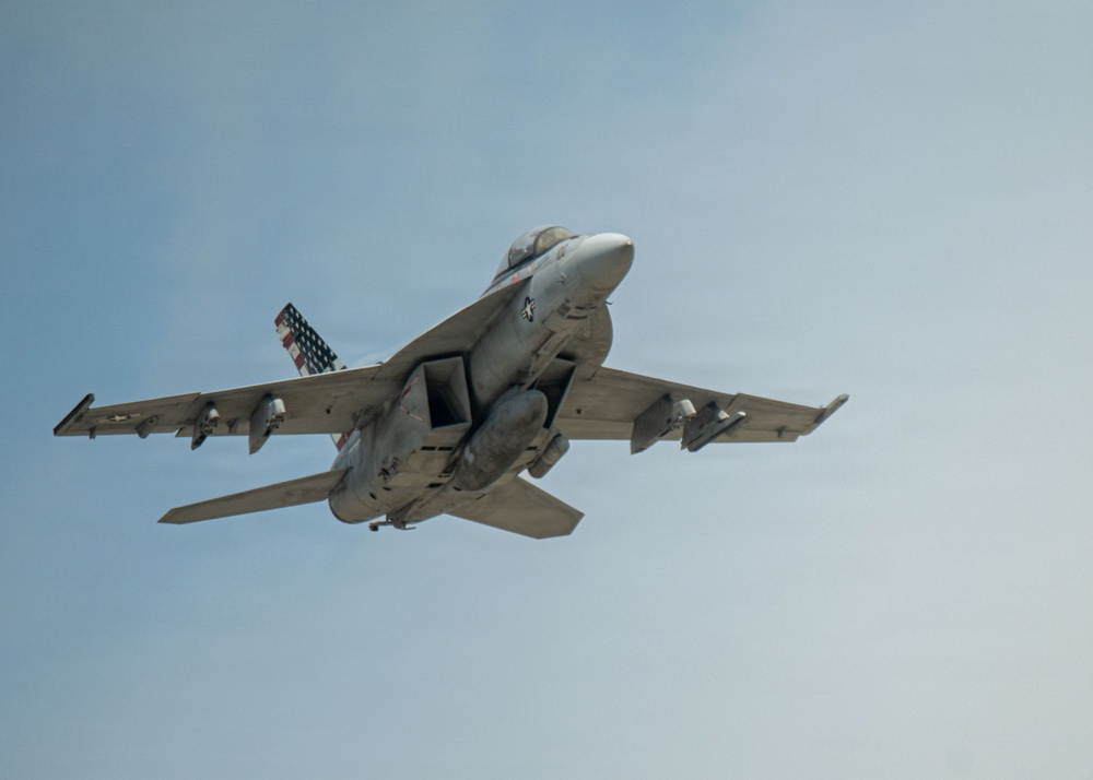 A U.S. Navy tactical jet aircraft, consistent with an F/A-18 Hornet or Super Hornet, is photographed from below at close range in flight against a pale blue sky. The aircraft is in a slight bank, revealing its underside with multiple hardpoints and ordnance or fuel tanks mounted. A distinctive American flag livery is visible on the tail fin.