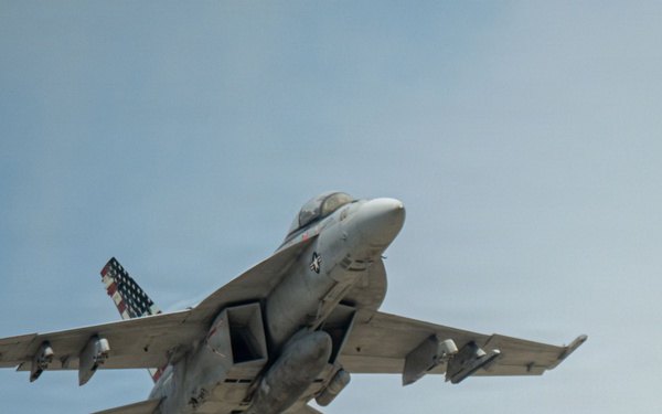 A U.S. Navy tactical jet aircraft, consistent with an F/A-18 Hornet or Super Hornet, is photographed from below at close range in flight against a pale blue sky. The aircraft is in a slight bank, revealing its underside with multiple hardpoints and ordnance or fuel tanks mounted. A distinctive American flag livery is visible on the tail fin.
