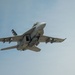 A U.S. Navy tactical jet aircraft, consistent with an F/A-18 Hornet or Super Hornet, is photographed from below at close range in flight against a pale blue sky. The aircraft is in a slight bank, revealing its underside with multiple hardpoints and ordnance or fuel tanks mounted. A distinctive American flag livery is visible on the tail fin.
