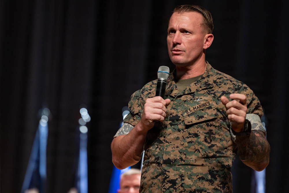 A U.S. Marines servicemember in digital camouflage uniform is speaking into a handheld microphone at what appears to be a formal event or briefing. He is gesturing with both hands in an engaged, expressive manner, suggesting he is delivering a speech or presentation. The background is dark with blurred bokeh lighting elements.