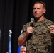 A U.S. Marines servicemember in digital camouflage uniform is speaking into a handheld microphone at what appears to be a formal event or briefing. He is gesturing with both hands in an engaged, expressive manner, suggesting he is delivering a speech or presentation. The background is dark with blurred bokeh lighting elements.
