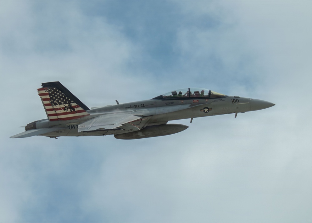 A U.S. Navy F/A-18 Super Hornet jet fighter is captured in flight against a partly cloudy sky. The aircraft bears the markings of VFA-32 (Strike Fighter Squadron 32) with modex number '100', indicating it is the Commanding Officer's aircraft. The tail features a distinctive American flag livery with what appears to be a commemorative or special paint scheme.