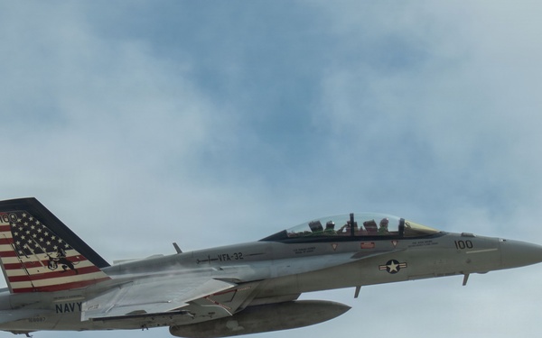 A U.S. Navy F/A-18 Super Hornet jet fighter is captured in flight against a partly cloudy sky. The aircraft bears the markings of VFA-32 (Strike Fighter Squadron 32) with modex number '100', indicating it is the Commanding Officer's aircraft. The tail features a distinctive American flag livery with what appears to be a commemorative or special paint scheme.