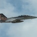 A U.S. Navy F/A-18 Super Hornet jet fighter is captured in flight against a partly cloudy sky. The aircraft bears the markings of VFA-32 (Strike Fighter Squadron 32) with modex number '100', indicating it is the Commanding Officer's aircraft. The tail features a distinctive American flag livery with what appears to be a commemorative or special paint scheme.