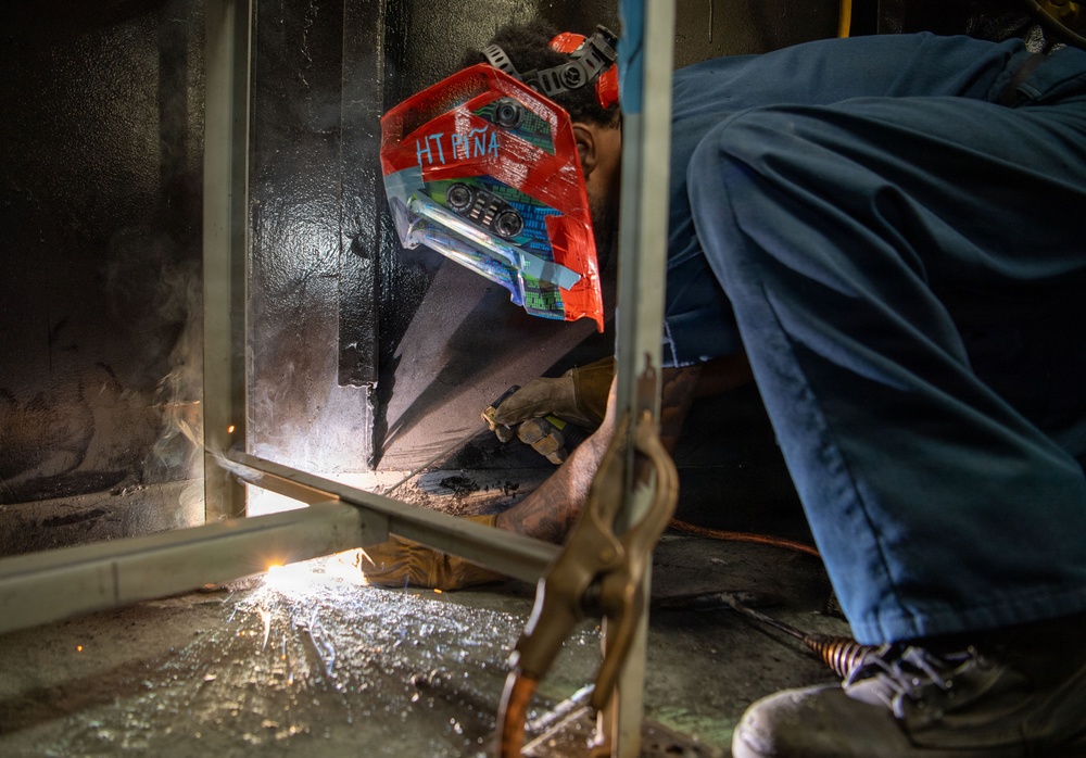 A welder is crouched low to the ground performing a welding operation at the base of a metal structure or panel. The worker is wearing a decorated red welding helmet with text reading 'HT PIÑA' and protective gloves, with visible sparks and light emanating from the weld point. The image captures an industrial work environment with smoke and scattered debris on the floor.