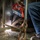 A welder is crouched low to the ground performing a welding operation at the base of a metal structure or panel. The worker is wearing a decorated red welding helmet with text reading 'HT PIÑA' and protective gloves, with visible sparks and light emanating from the weld point. The image captures an industrial work environment with smoke and scattered debris on the floor.