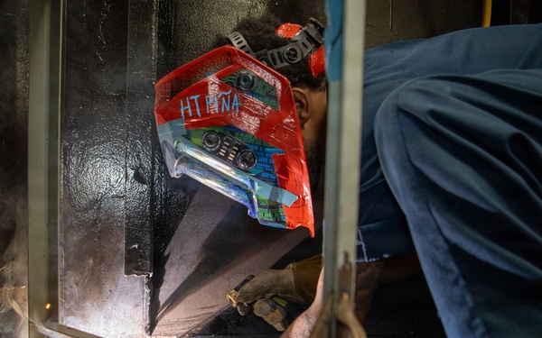 A welder is crouched low to the ground performing a welding operation at the base of a metal structure or panel. The worker is wearing a decorated red welding helmet with text reading 'HT PIÑA' and protective gloves, with visible sparks and light emanating from the weld point. The image captures an industrial work environment with smoke and scattered debris on the floor.