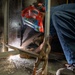 A welder is crouched low to the ground performing a welding operation at the base of a metal structure or panel. The worker is wearing a decorated red welding helmet with text reading 'HT PIÑA' and protective gloves, with visible sparks and light emanating from the weld point. The image captures an industrial work environment with smoke and scattered debris on the floor.