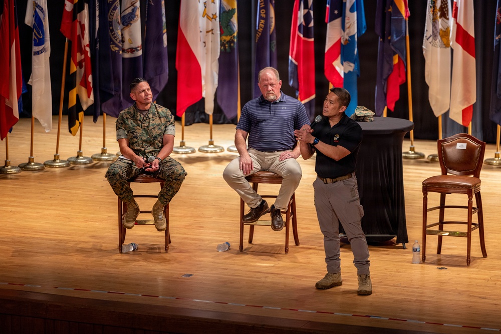 Three individuals are on a wooden stage during what appears to be a panel discussion or speaking event at a military venue. Two men are seated on bar-height chairs while a third stands holding a microphone and speaking. The backdrop features numerous U.S. state and military flags arranged in a row.