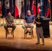 Three individuals are on a wooden stage during what appears to be a panel discussion or speaking event at a military venue. Two men are seated on bar-height chairs while a third stands holding a microphone and speaking. The backdrop features numerous U.S. state and military flags arranged in a row.
