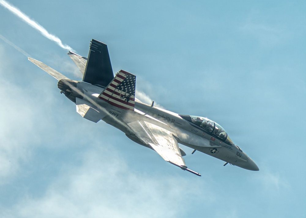 A U.S. Navy F/A-18 Super Hornet from VFA-32 (Fighting Swordsmen) is captured in a steep banking maneuver against a partly cloudy sky. The aircraft bears the modex number '100' and features a distinctive American flag livery on its vertical stabilizer. A vapor contrail is visible trailing from the aircraft, indicating high-speed or high-G flight conditions.