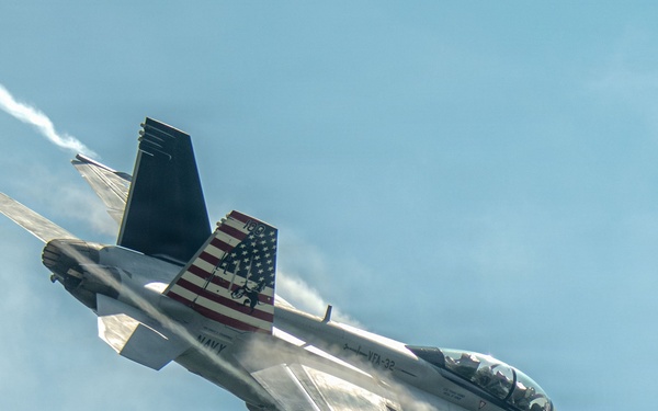 A U.S. Navy F/A-18 Super Hornet from VFA-32 (Fighting Swordsmen) is captured in a steep banking maneuver against a partly cloudy sky. The aircraft bears the modex number '100' and features a distinctive American flag livery on its vertical stabilizer. A vapor contrail is visible trailing from the aircraft, indicating high-speed or high-G flight conditions.