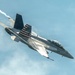 A U.S. Navy F/A-18 Super Hornet from VFA-32 (Fighting Swordsmen) is captured in a steep banking maneuver against a partly cloudy sky. The aircraft bears the modex number '100' and features a distinctive American flag livery on its vertical stabilizer. A vapor contrail is visible trailing from the aircraft, indicating high-speed or high-G flight conditions.