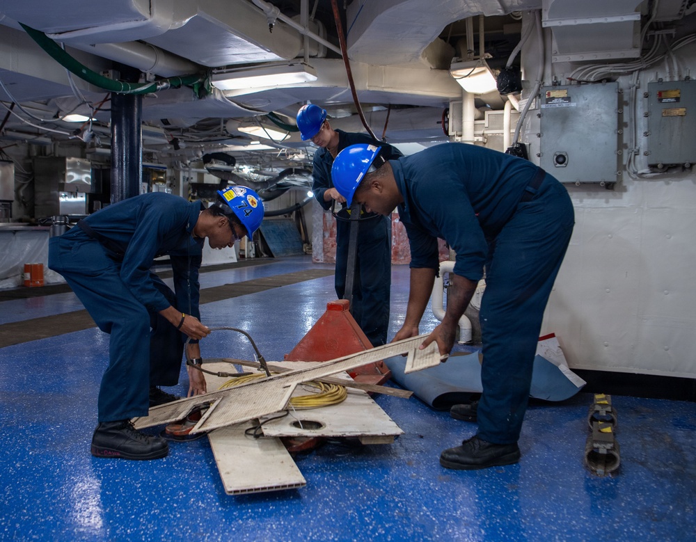 Three personnel wearing blue coveralls and blue hard hats are working on the hangar deck of what appears to be a U.S. Navy aircraft carrier. They are handling and sorting through various materials including boards, cables, and equipment on a blue-painted deck. The interior space shows typical naval vessel infrastructure including overhead pipes, conduits, and electrical panels.