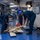 Three personnel wearing blue coveralls and blue hard hats are working on the hangar deck of what appears to be a U.S. Navy aircraft carrier. They are handling and sorting through various materials including boards, cables, and equipment on a blue-painted deck. The interior space shows typical naval vessel infrastructure including overhead pipes, conduits, and electrical panels.