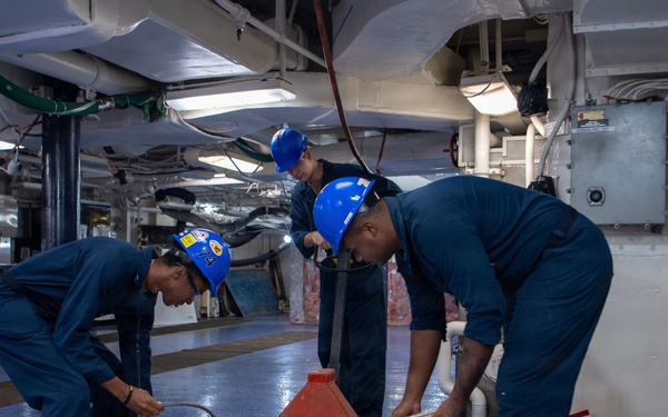 Three personnel wearing blue coveralls and blue hard hats are working on the hangar deck of what appears to be a U.S. Navy aircraft carrier. They are handling and sorting through various materials including boards, cables, and equipment on a blue-painted deck. The interior space shows typical naval vessel infrastructure including overhead pipes, conduits, and electrical panels.