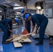 Three personnel wearing blue coveralls and blue hard hats are working on the hangar deck of what appears to be a U.S. Navy aircraft carrier. They are handling and sorting through various materials including boards, cables, and equipment on a blue-painted deck. The interior space shows typical naval vessel infrastructure including overhead pipes, conduits, and electrical panels.