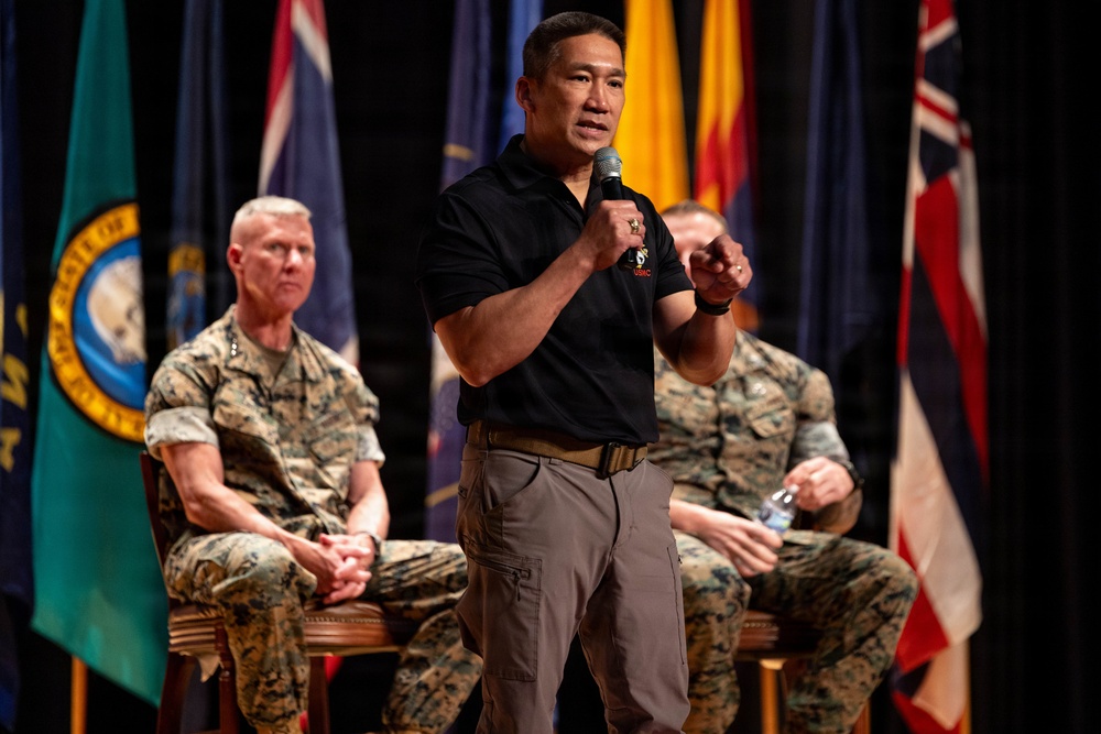 A man in civilian attire (black polo shirt and gray cargo pants) speaks into a microphone on a stage, gesturing with his right hand. Behind him, two military personnel in Marine Corps MARPAT camouflage uniforms are seated in chairs. Multiple flags, including what appear to be U.S. state flags and possibly the Australian flag, are displayed in the background.