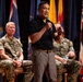 A man in civilian attire (black polo shirt and gray cargo pants) speaks into a microphone on a stage, gesturing with his right hand. Behind him, two military personnel in Marine Corps MARPAT camouflage uniforms are seated in chairs. Multiple flags, including what appear to be U.S. state flags and possibly the Australian flag, are displayed in the background.