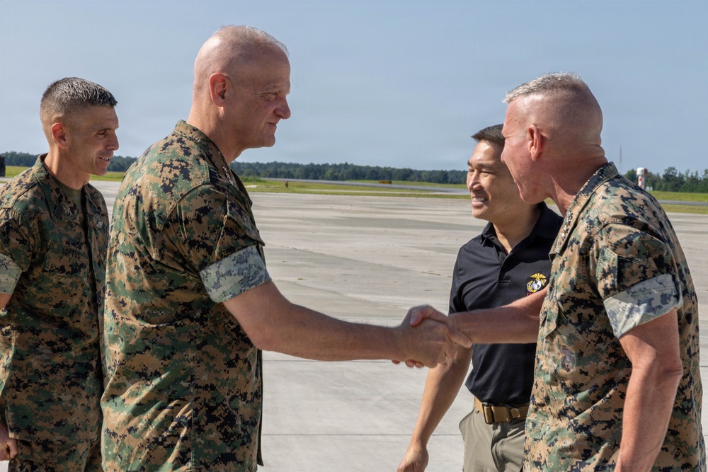 Four individuals are gathered on a military airfield tarmac, with two U.S. Marines in MARPAT camouflage uniforms shaking hands in a formal greeting. A civilian in a black polo shirt with an emblem and another Marine in uniform stand nearby observing the exchange. The setting appears to be an official arrival or greeting ceremony at a military air station.