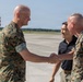 Four individuals are gathered on a military airfield tarmac, with two U.S. Marines in MARPAT camouflage uniforms shaking hands in a formal greeting. A civilian in a black polo shirt with an emblem and another Marine in uniform stand nearby observing the exchange. The setting appears to be an official arrival or greeting ceremony at a military air station.