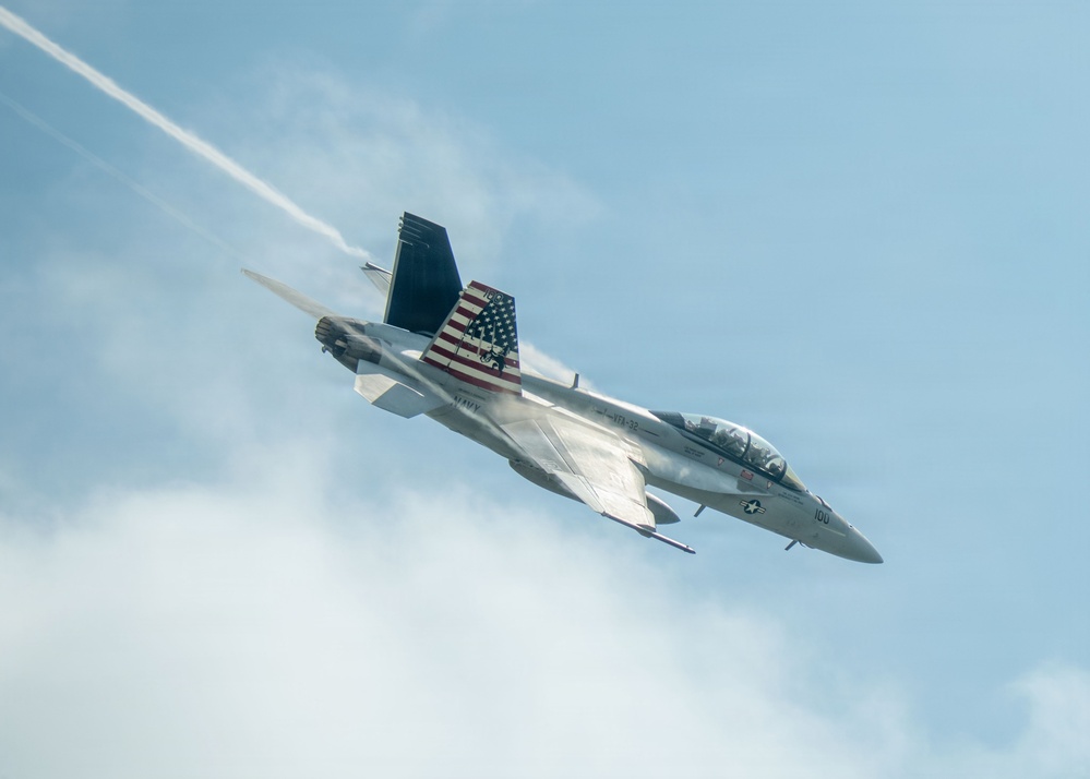 A U.S. Navy F/A-18 Super Hornet jet fighter is captured in a steep banking maneuver against a partly cloudy blue sky. The aircraft bears the number '100' on its nose and features a prominent American flag livery on its tail fin, along with 'NAVY' markings on the fuselage. A white vapor/condensation trail is visible streaming from the aircraft, likely from wingtip vortices or engine exhaust during the high-performance maneuver.