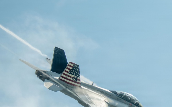 A U.S. Navy F/A-18 Super Hornet jet fighter is captured in a steep banking maneuver against a partly cloudy blue sky. The aircraft bears the number '100' on its nose and features a prominent American flag livery on its tail fin, along with 'NAVY' markings on the fuselage. A white vapor/condensation trail is visible streaming from the aircraft, likely from wingtip vortices or engine exhaust during the high-performance maneuver.
