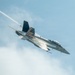 A U.S. Navy F/A-18 Super Hornet jet fighter is captured in a steep banking maneuver against a partly cloudy blue sky. The aircraft bears the number '100' on its nose and features a prominent American flag livery on its tail fin, along with 'NAVY' markings on the fuselage. A white vapor/condensation trail is visible streaming from the aircraft, likely from wingtip vortices or engine exhaust during the high-performance maneuver.