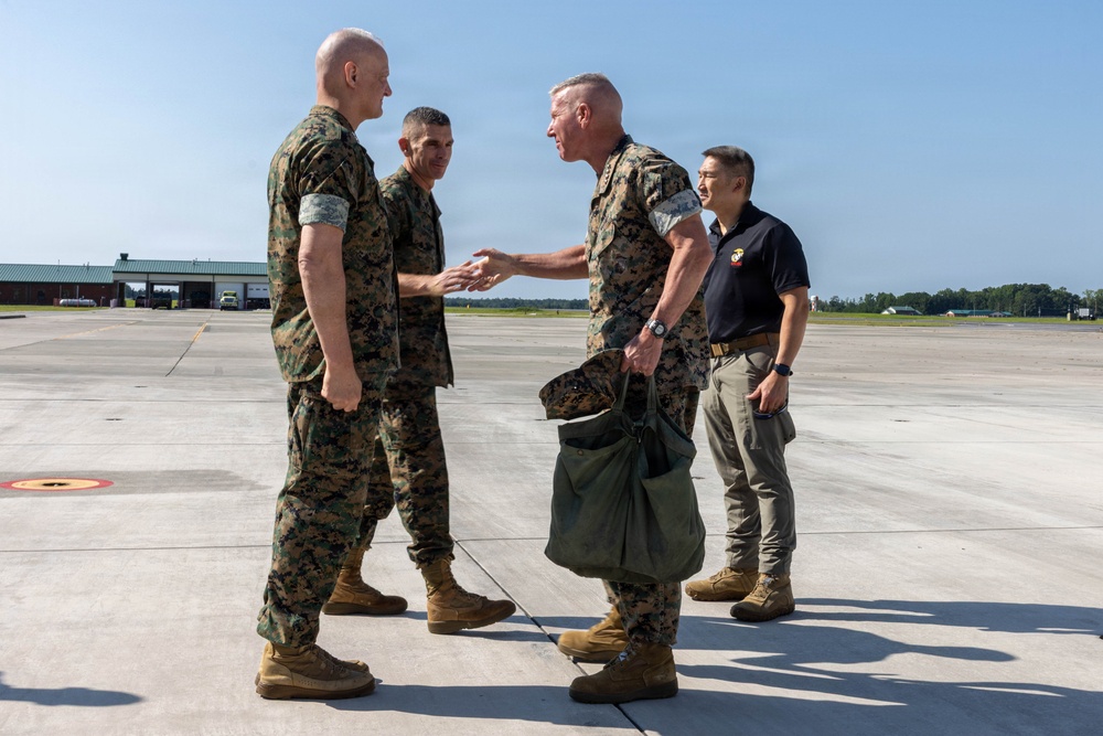 Four individuals are gathered on a military airfield tarmac, with three wearing U.S. Marine Corps MARPAT camouflage uniforms and one in civilian attire. Two Marines appear to be engaged in a greeting or handshake, suggesting an arrival or reception ceremony. The setting appears to be a military air station during daytime with clear skies.