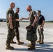 Four individuals are gathered on a military airfield tarmac, with three wearing U.S. Marine Corps MARPAT camouflage uniforms and one in civilian attire. Two Marines appear to be engaged in a greeting or handshake, suggesting an arrival or reception ceremony. The setting appears to be a military air station during daytime with clear skies.